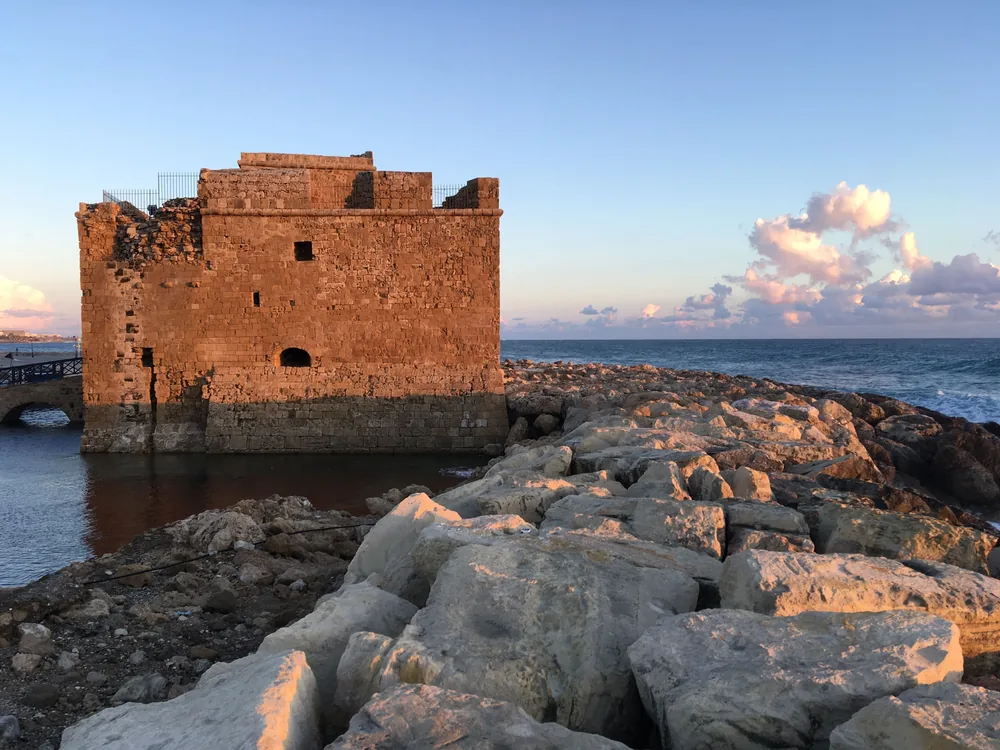 Paphos Castle, a square stone fort at the water's edge, glowing gold at sunset beside a rocky breakwater