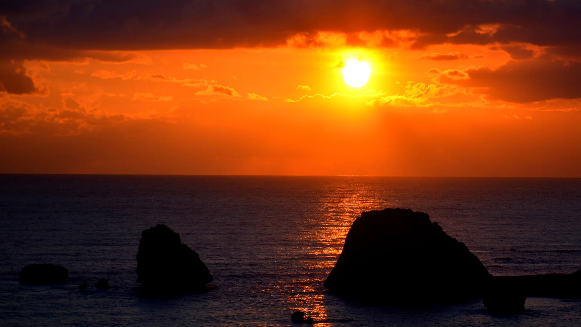 The sun setting through an orange sky over a calm sea, with dark sea stacks silhouetted in the water