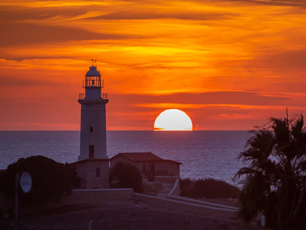 A white lighthouse silhouetted against a deep orange sky as the sun touches the sea
